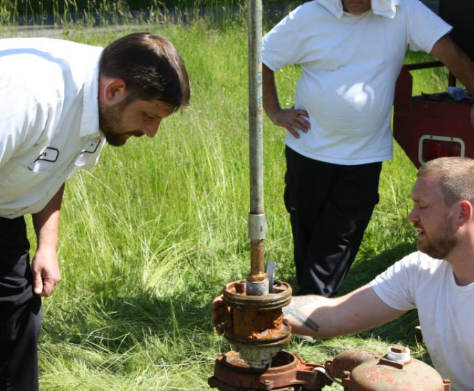 Three men examining rusty machinery outdoors; one pointing, others observing. Green grass in background.