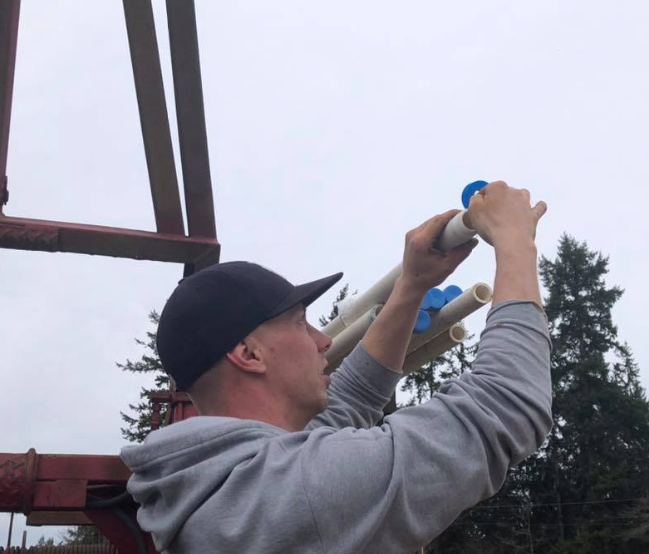 Man in a gray hoodie and cap assembling PVC pipes outdoors, against a cloudy sky.