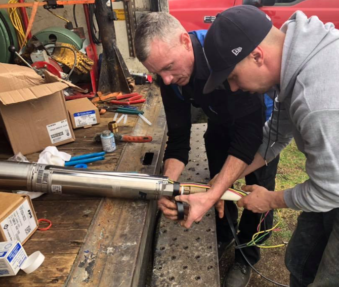 Two workers assemble a cylindrical water pump with tools on a truck bed.