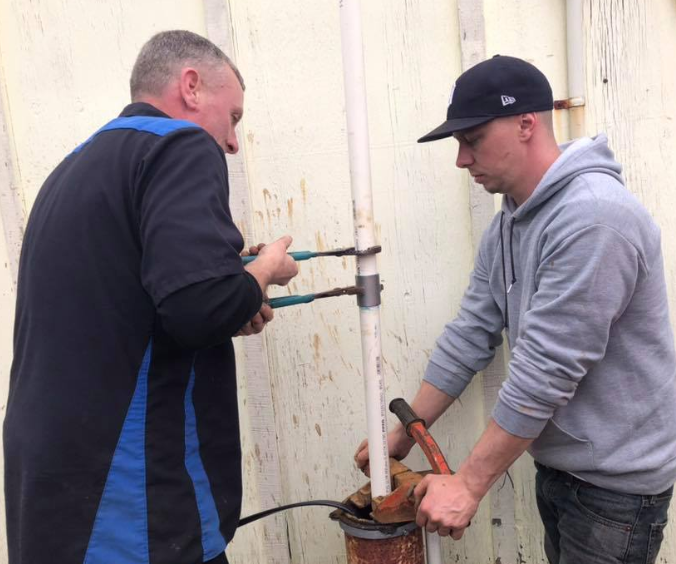 Two men working on a pipe with tools. One uses pliers while the other operates a pump outdoors.