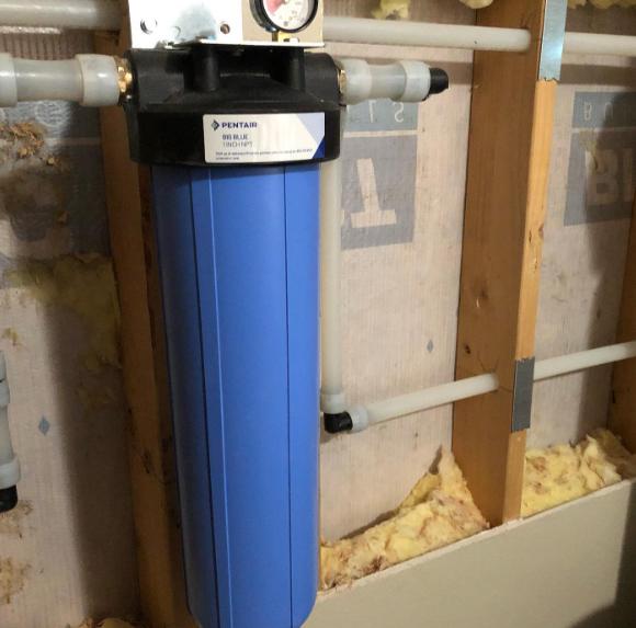 Blue water filter system installed on a wall with white PEX pipes and pressure gauge.