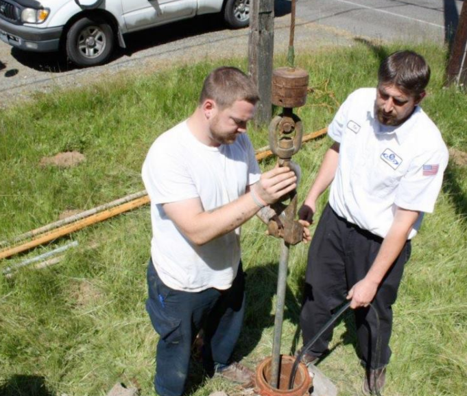 Two men working on a well in a grassy area, one in a white shirt and the other in a work shirt and dark pants.