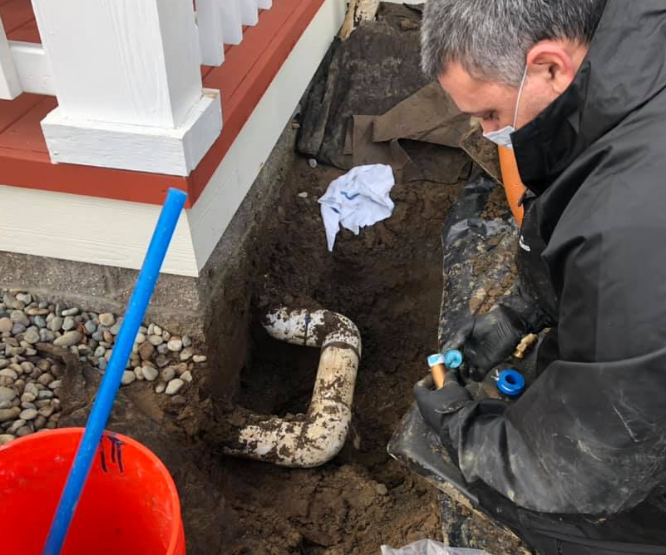 Man working on a broken pipe in a trench next to a house. The man wears a mask.
