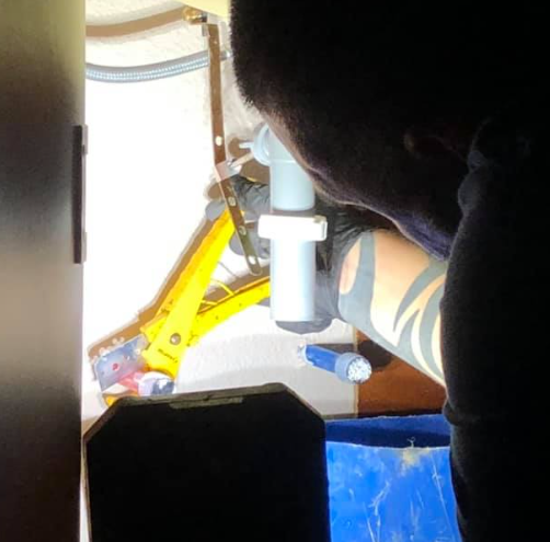 Person with arm tattoos working on plumbing under a sink, using a yellow jack, with a flashlight.