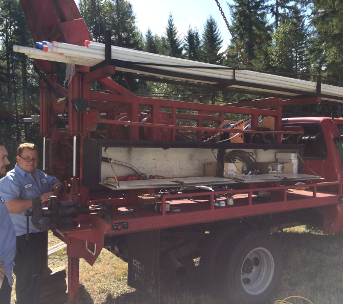 Red well-drilling truck with two men in blue shirts, outdoors. Pipes are on top of the truck, trees in the background.