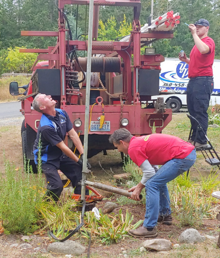 Men working on a well with a red truck, one looking up, others handling equipment outdoors.