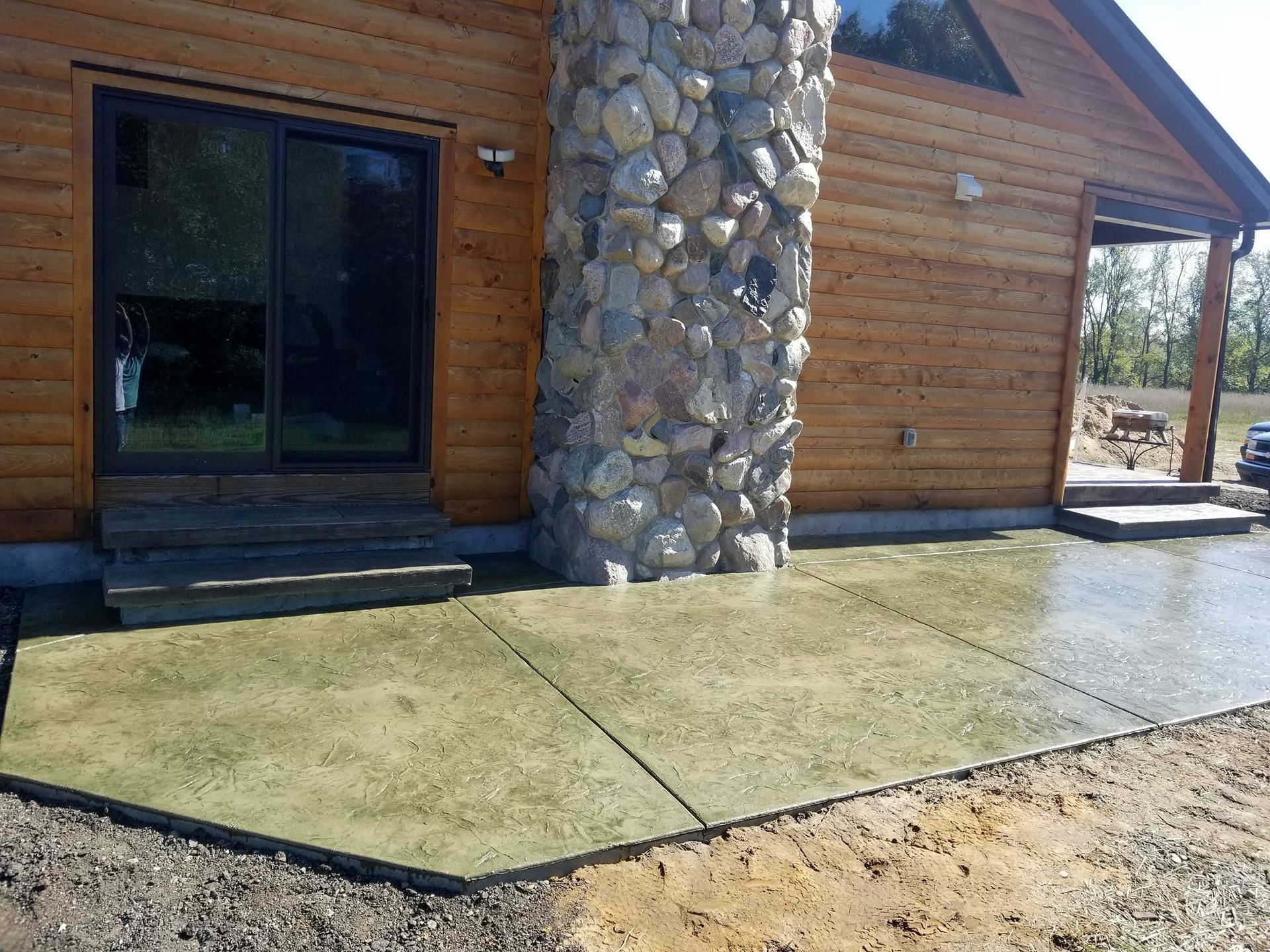 A log cabin with a stone chimney and a concrete patio in front of it.