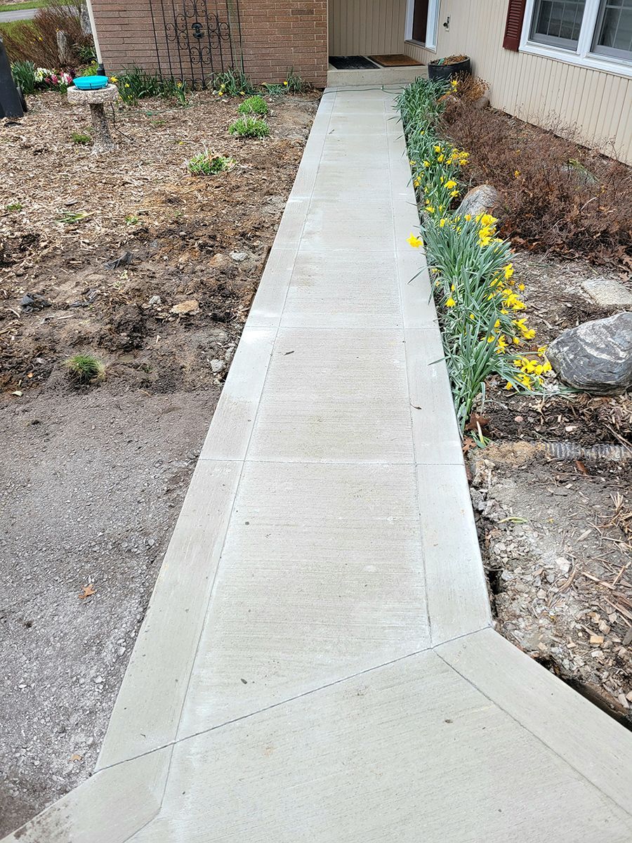 A concrete walkway leading to a house with yellow flowers on the side.