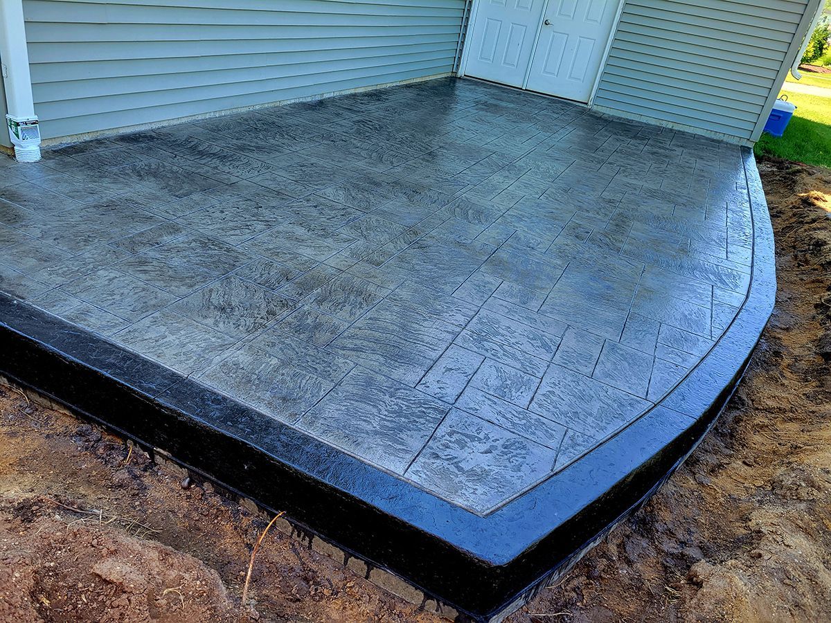 A concrete patio with a black border is sitting in the dirt in front of a house.