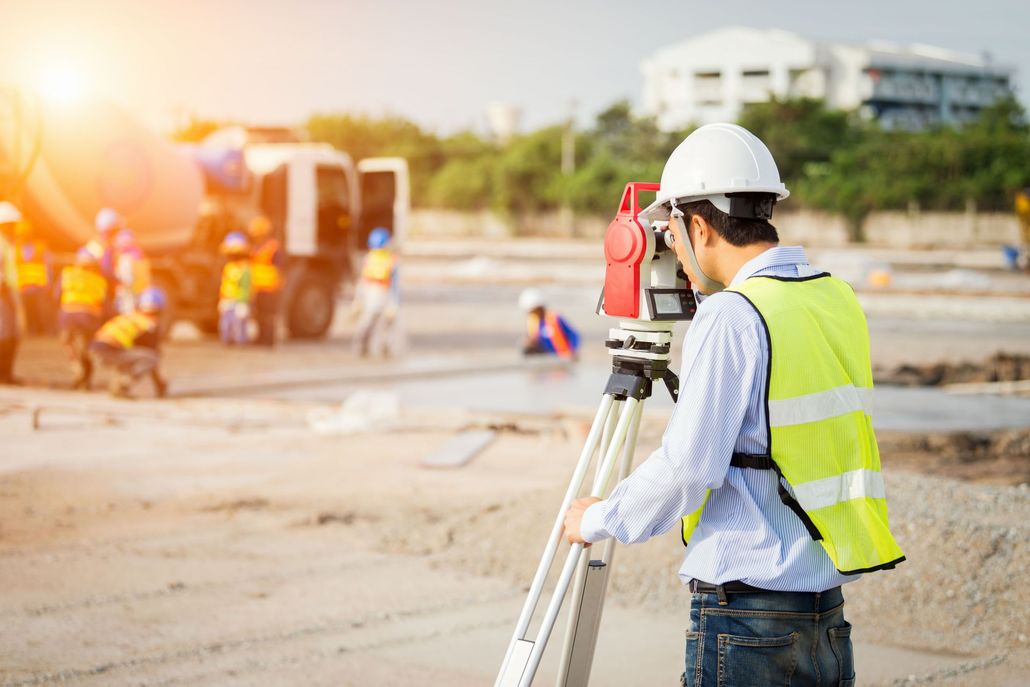 Surveyor in white hard hat and yellow vest using a tripod instrument at a construction site with workers in background