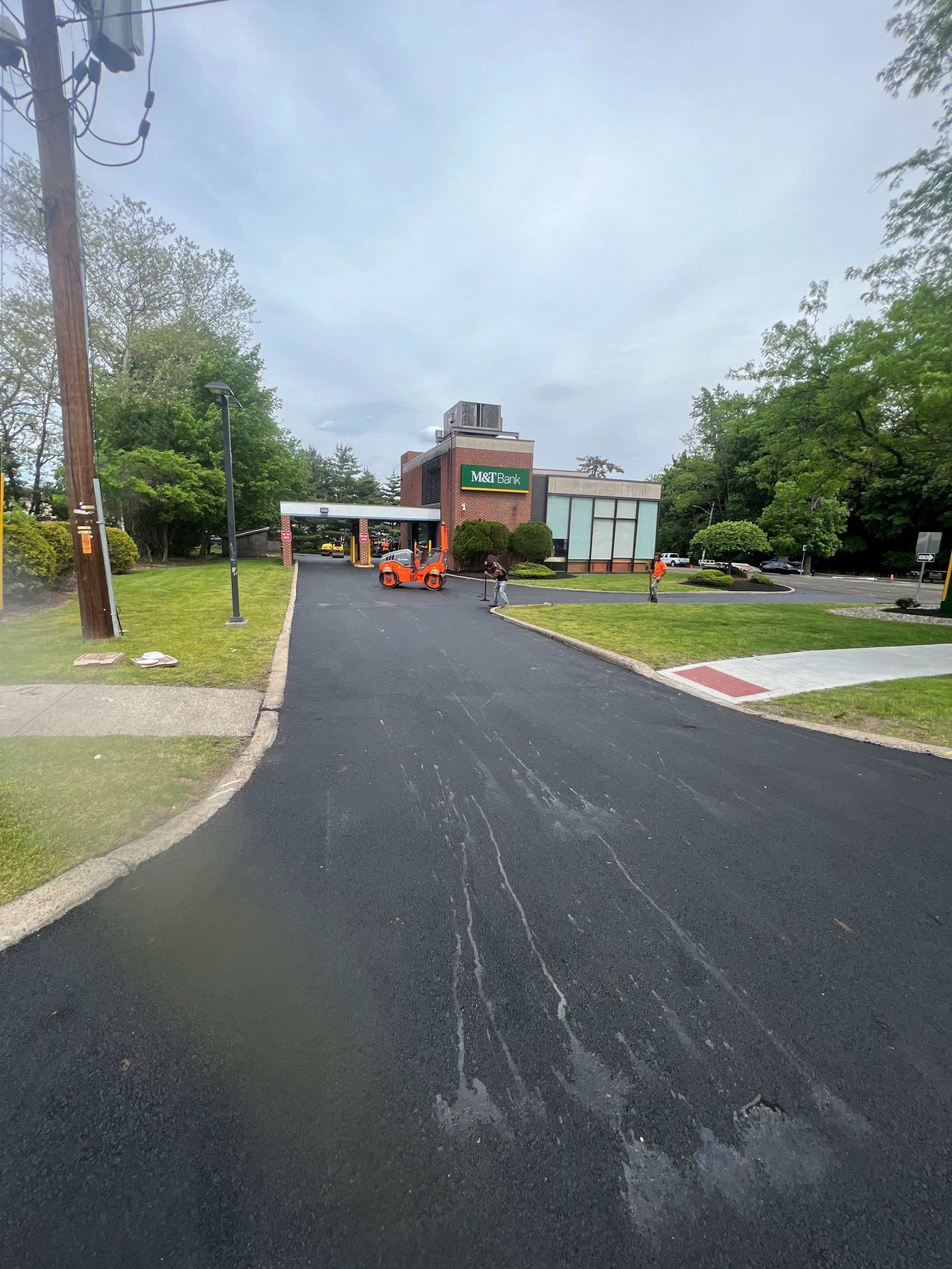 New asphalt driveway leading to a brick building, with an orange lift and trees on both sides.
