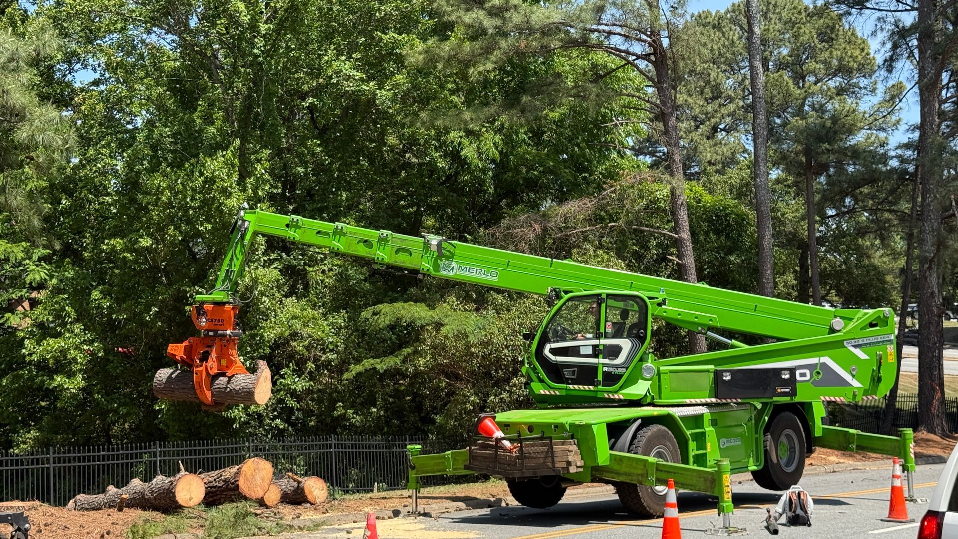 Green boom truck lifting a worker basket beside cut logs in a wooded roadside area