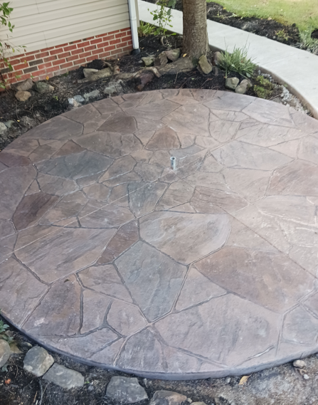 Circular stone patio beside a house, bordered by dark rocks and a curved walkway.