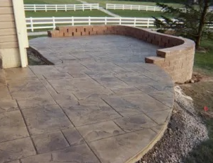 Curved stone patio with stamped concrete surface and low brick seating wall beside a house
