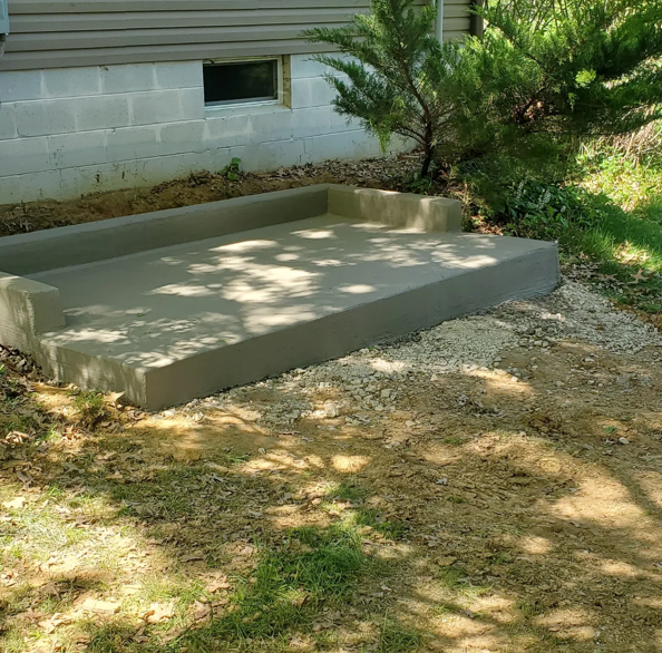 Concrete patio slab beside a house, bordered by grass and a bush in a shaded yard.