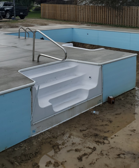 Broken pool entrance with a white molded wheelchair ramp and blue walls beside a drained swimming pool.