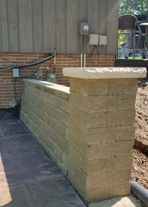 Partially built tan block retaining wall beside a house exterior and concrete patio