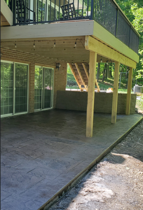 Covered patio under a raised deck with stone flooring, wooden posts, and sliding glass doors.