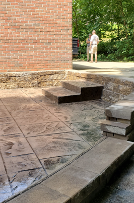 Brick building entrance with concrete steps, patterned patio, and a person standing in the background.