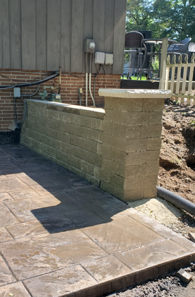 Stone patio and tan brick retaining wall beside a house, with a small yard and fence in the background