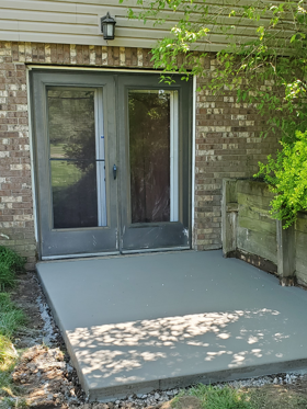 Concrete patio leading to gray double doors on a brick house, with a wall lantern and greenery nearby