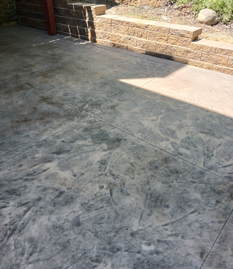 Concrete patio with sunlight and shadow beside a low stone retaining wall and garden bed