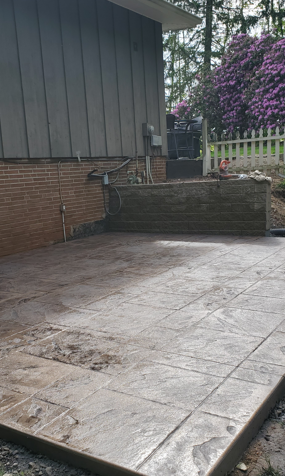Wet stone patio beside house with brick wall, steps, and flowering hedge in the background