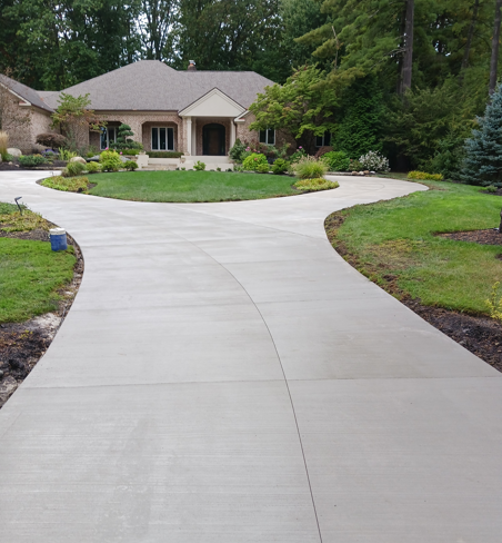 Curved concrete driveway leading to a house with landscaped lawn and trees