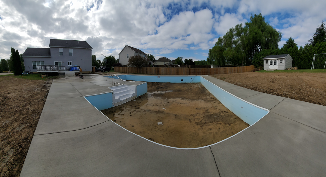 Empty concrete pool under construction in a suburban backyard, with blue lining and surrounding houses.