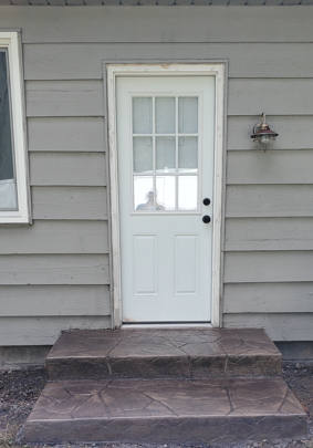Front door with white door and sidelights, set in gray siding above a small stone stoop.