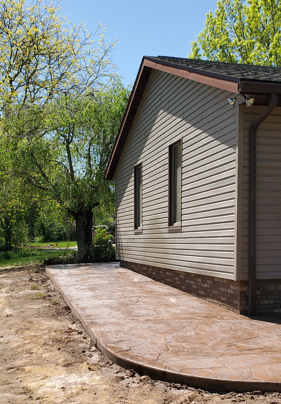 Side of a beige house with a curved concrete patio beside a wooded yard