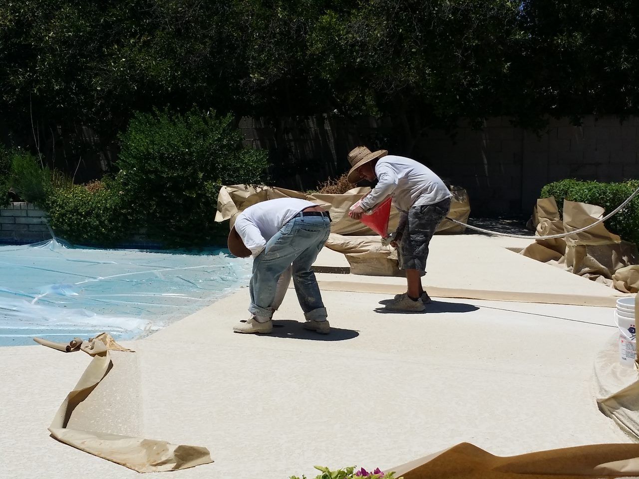 Two workers applying a light-colored coating around a swimming pool.