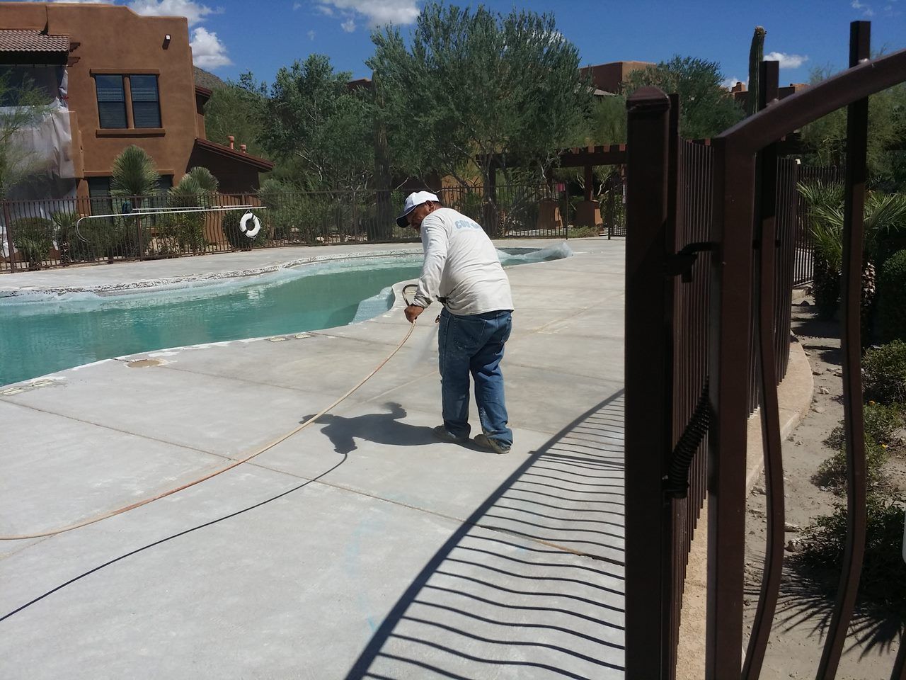 Person sprays near a pool. They wear a hat and jeans, in a sunny outdoor setting.