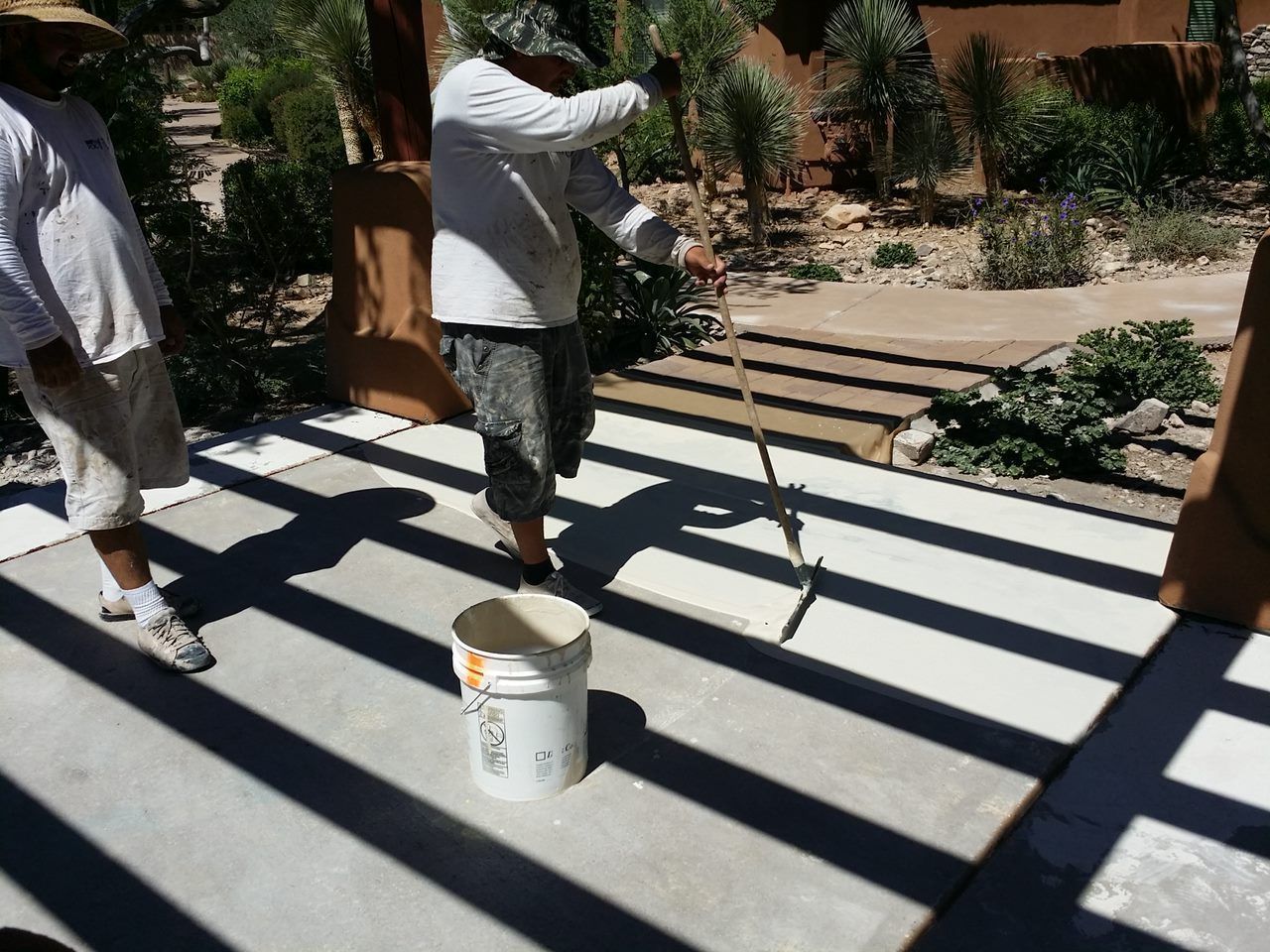 Workers applying coating to concrete patio outdoors; bucket and tool visible.