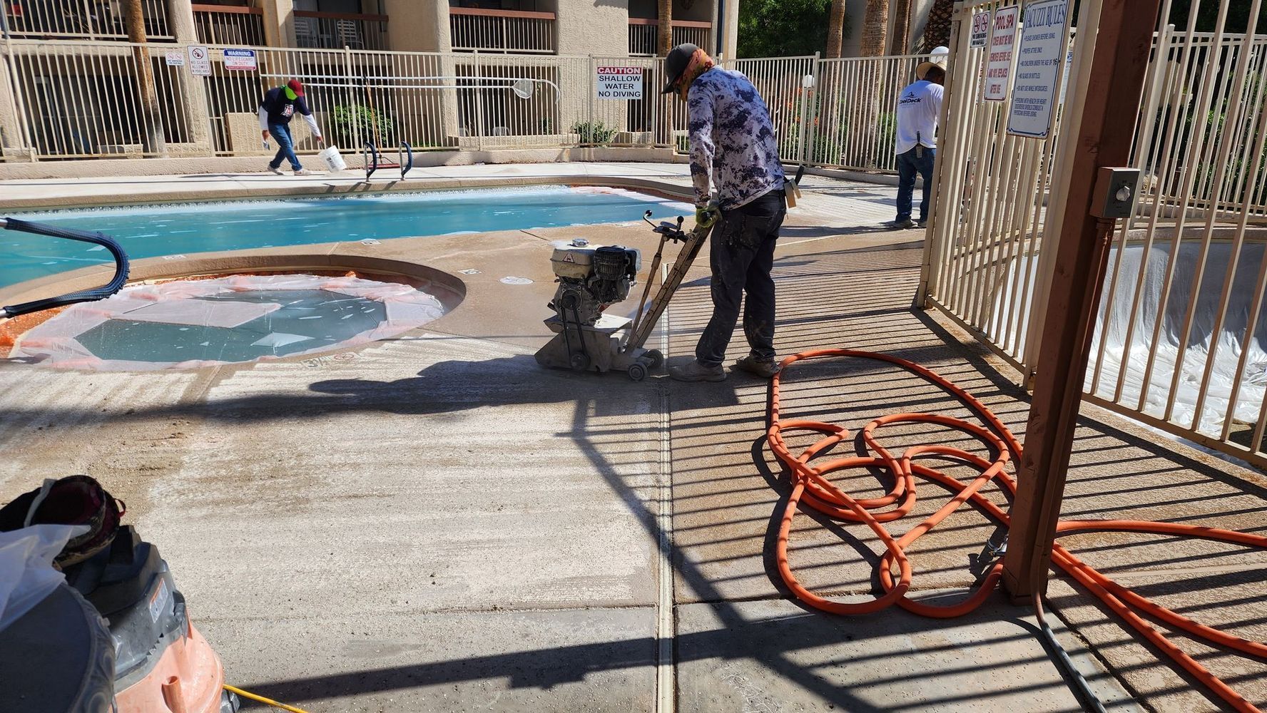 Man using a power tool to work on concrete near a pool. Orange hose and fence in the foreground.