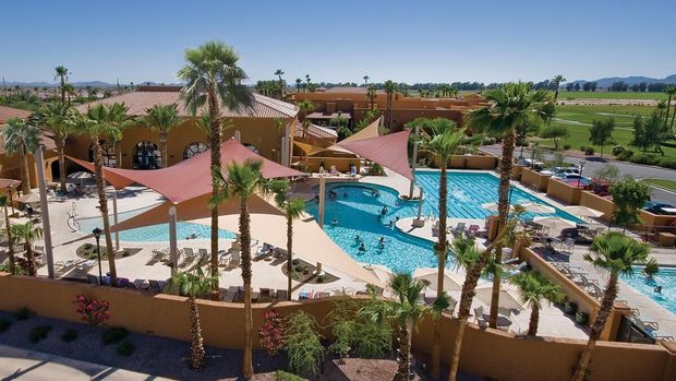 Pool area with shade sails, palm trees, and tan buildings under a blue sky.