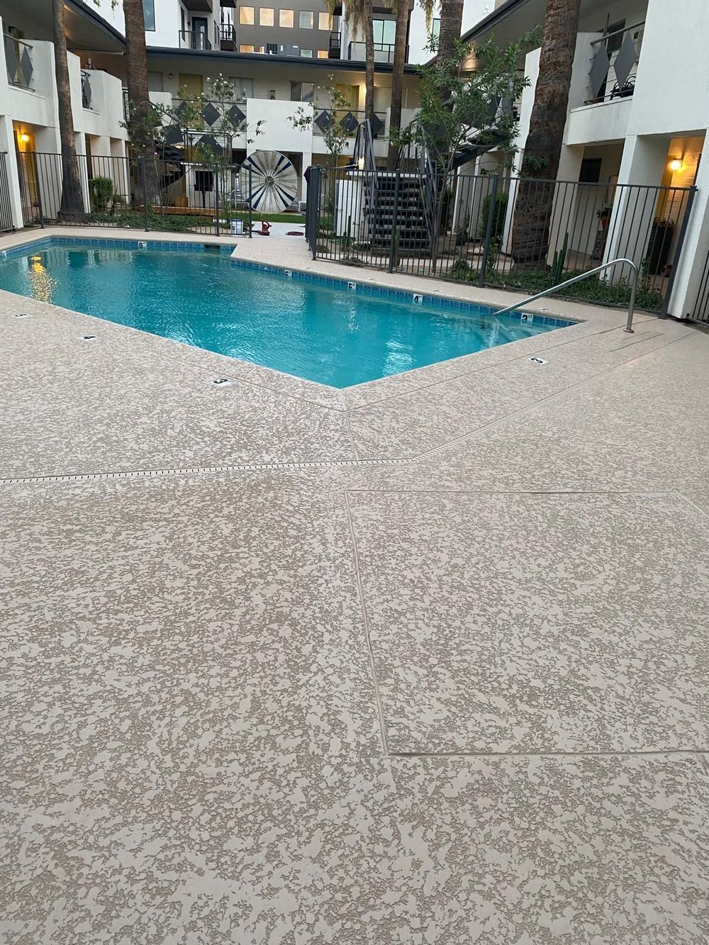 Pool with turquoise water surrounded by textured beige concrete. Apartment buildings and palm trees are visible.