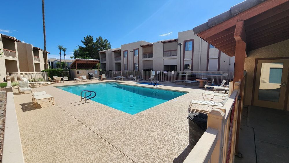 Pool area with turquoise water, beige paving, and buildings in the background under a blue sky.
