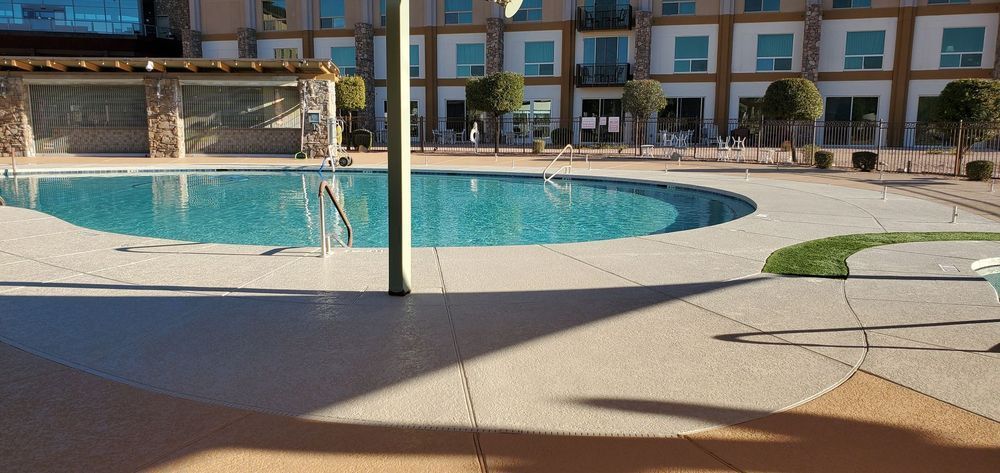 A pool with blue water and surrounding concrete, trees, and buildings. Sunny day.