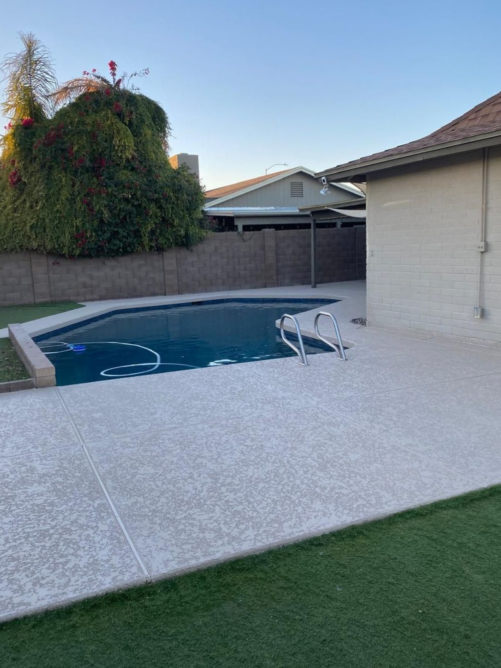 Backyard with a pool, concrete patio, green grass, and a beige stucco wall under a clear sky.