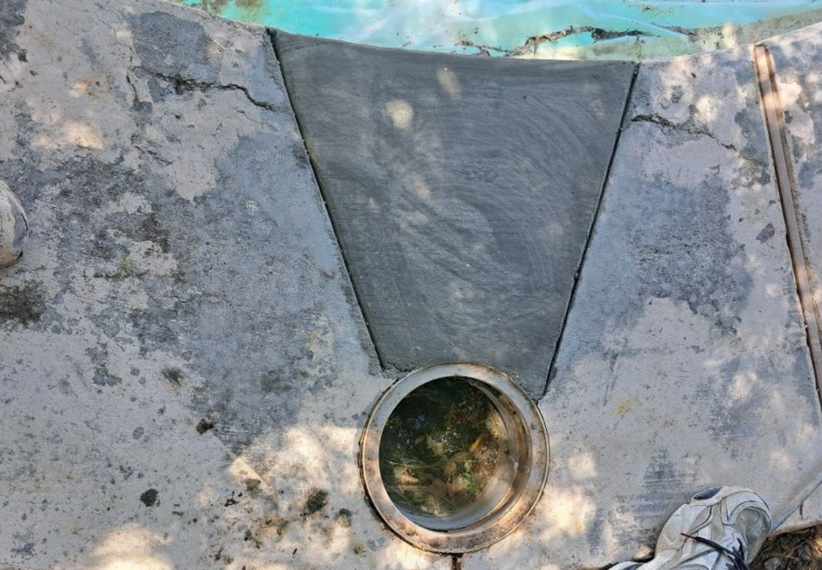 Concrete pool deck with open drain and partial view of pool water. A person stands nearby.