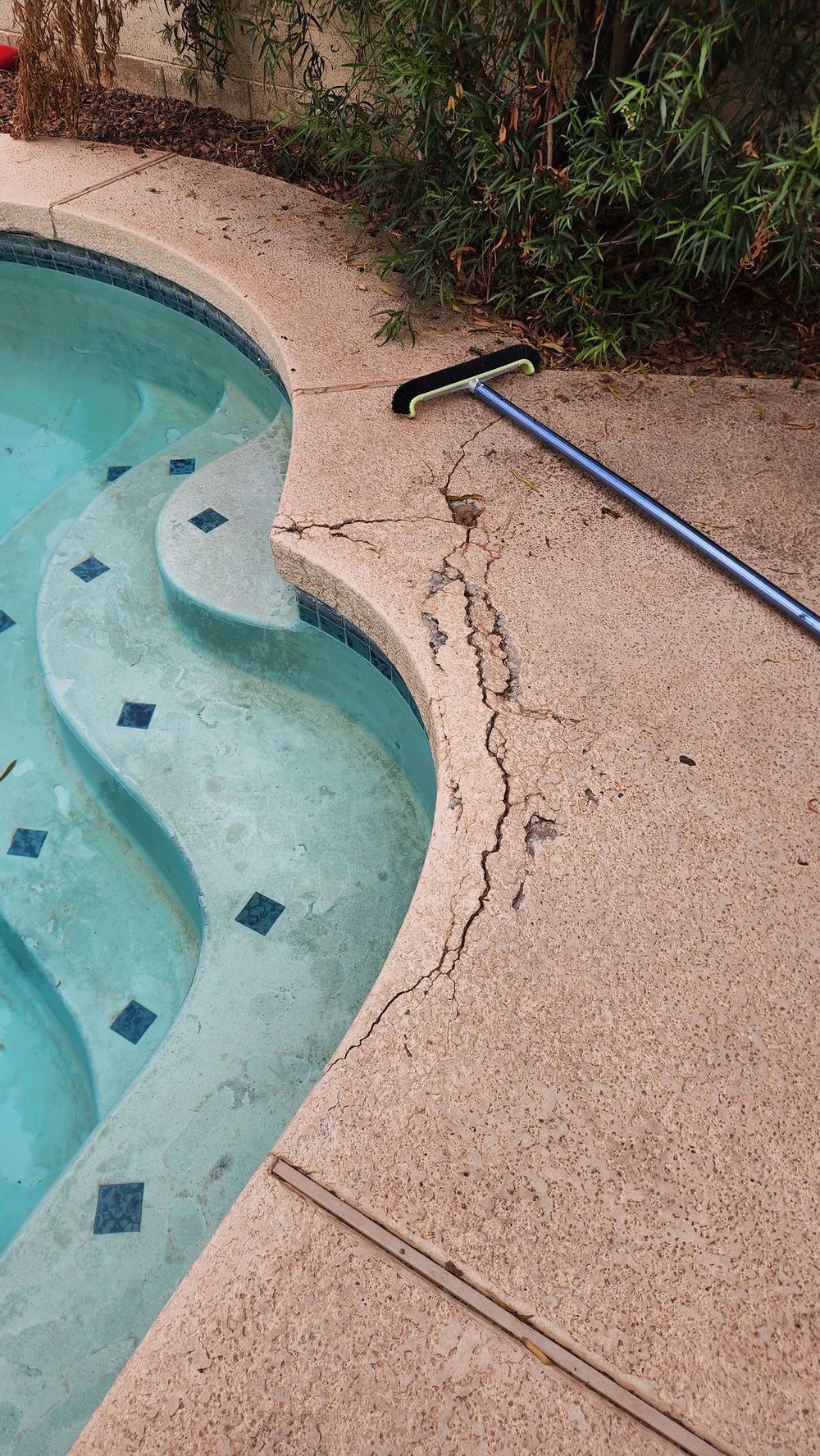 Cracked concrete pool deck next to a pool with steps, a black and blue pool brush rests on the deck.