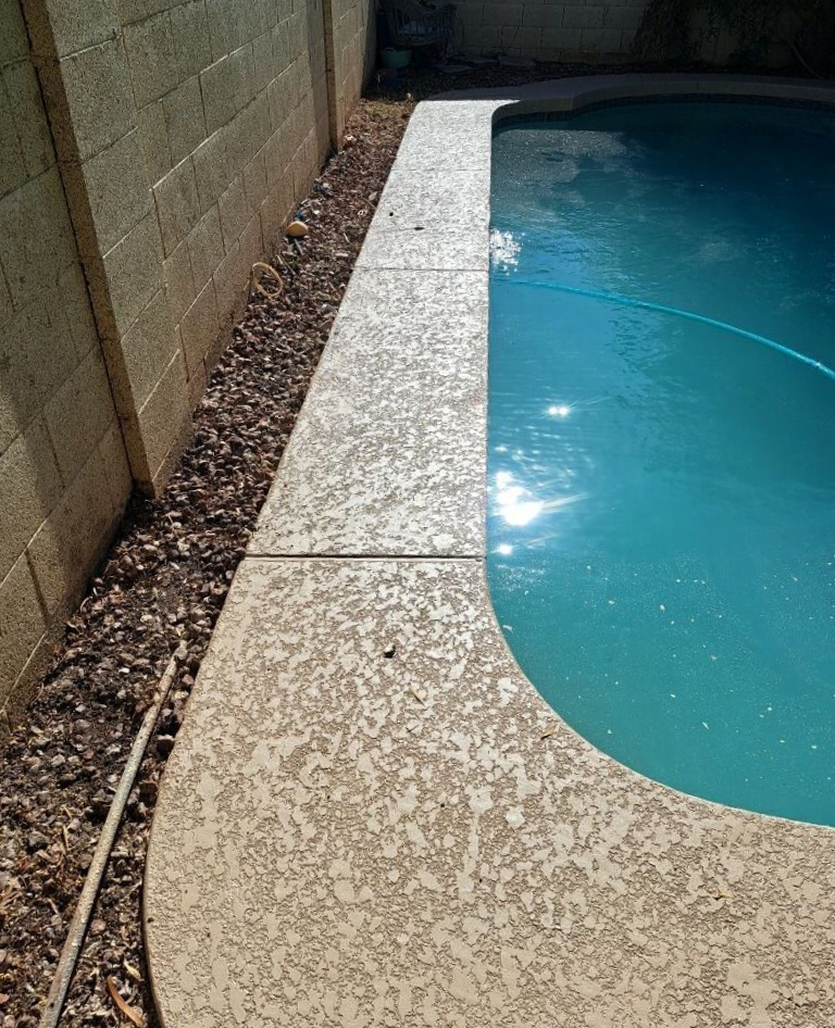 Poolside with a concrete walkway, pool, and a beige block wall. Small rocks line the walkway's edge.