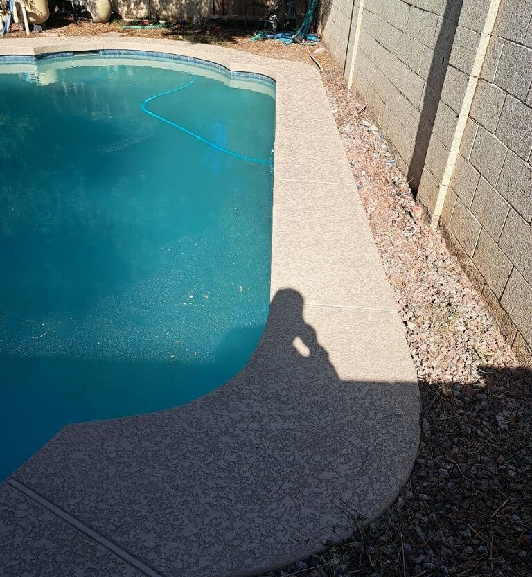 Poolside view: blue pool water, speckled concrete edge, gray wall, gravel, sunny day.