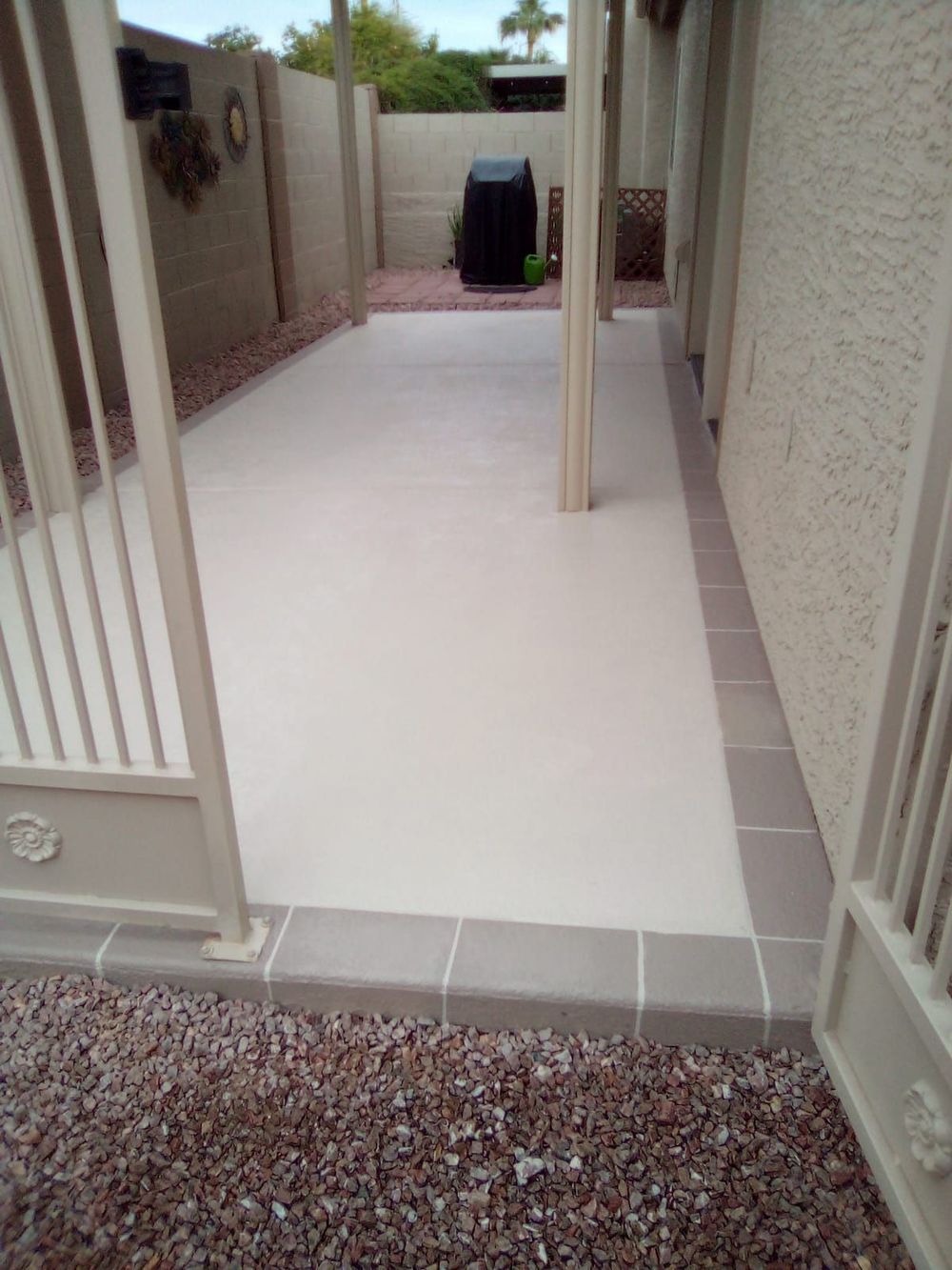 Concrete patio with gray border, adjacent to a textured beige wall and gated entrance with gravel ground.