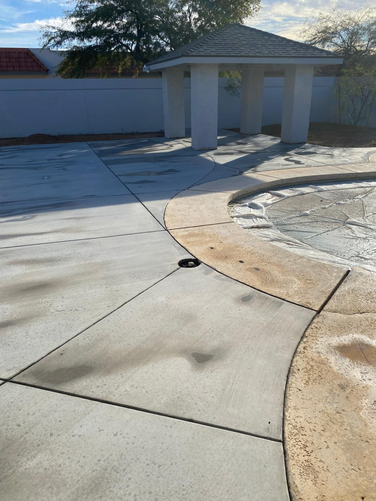 Concrete patio with a gazebo, connected to a pool with brown borders, and a white wall in the background.