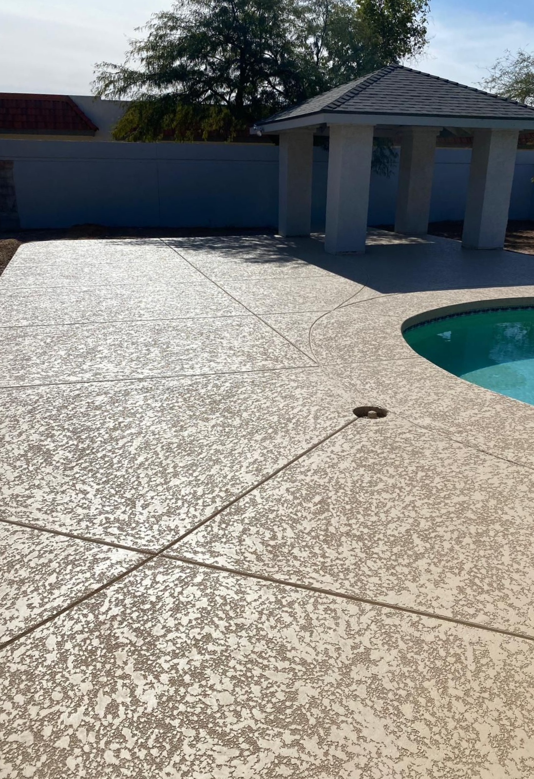 Concrete patio surrounding a pool and gazebo; textured surface in light beige.