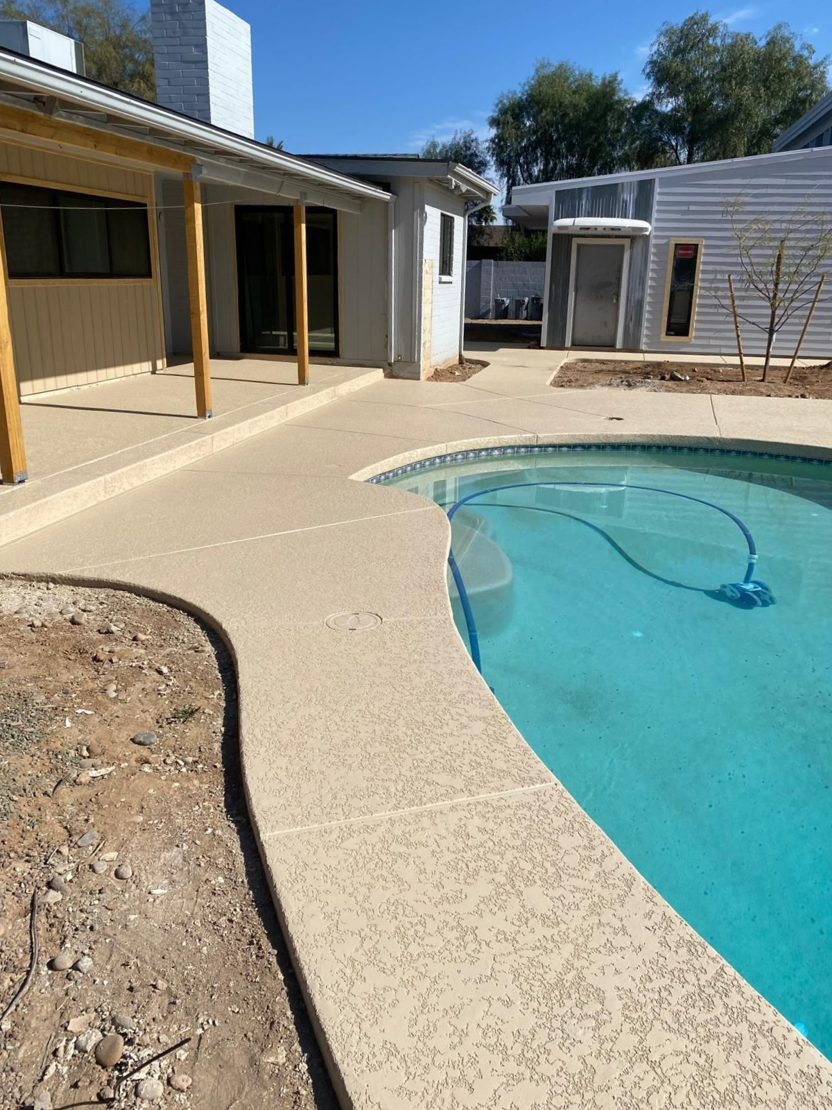 Backyard with a pool and concrete patio, next to a covered porch and a white building.