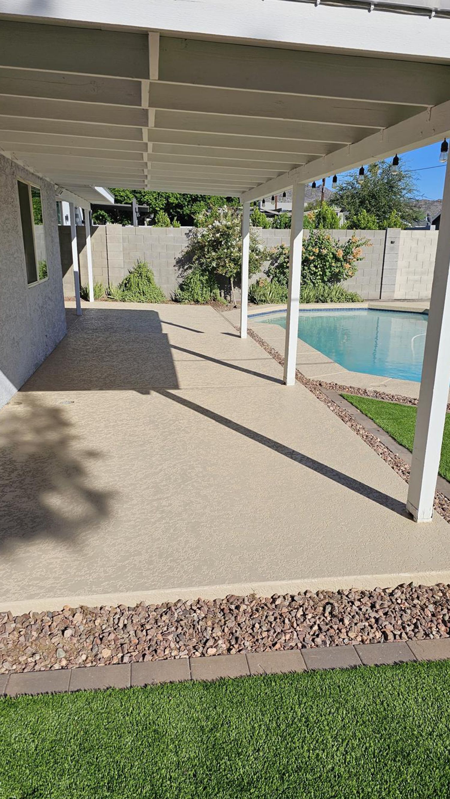 Patio with pergola, concrete flooring, and swimming pool bordered by grass and rocks.