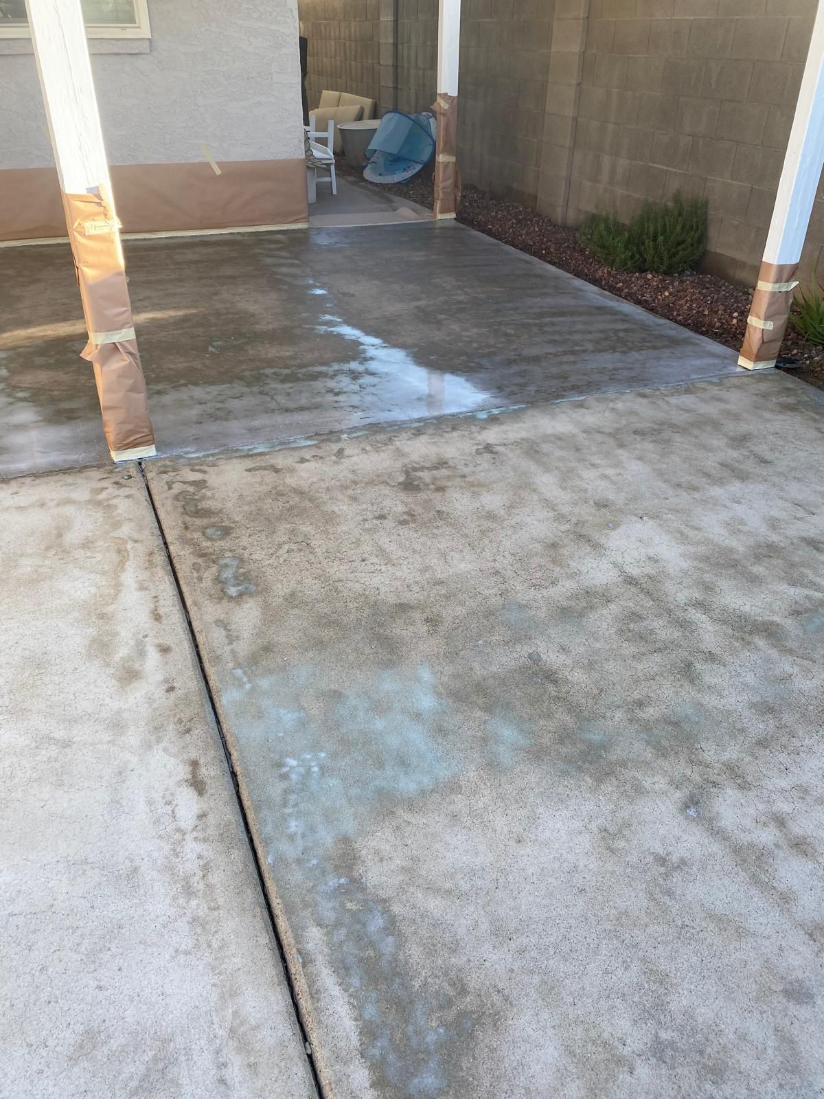 Concrete driveway under a covered area, showing a wet, textured surface with brown columns and a section of landscaping.